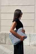 A woman in a black dress and sunglasses holds a chunky light blue and gray crocheted clutch bag against a beige stone wall background.