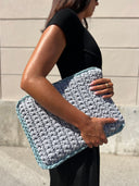 A woman in a black dress holds a large, chunky crocheted clutch bag in light gray with a light blue border, standing against a beige stone wall.