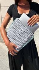 A person wearing a black dress holds a light gray chunky-knit laptop sleeve with a light blue edge, inserting a silver laptop into it.