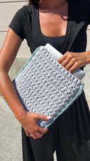 A person outdoors holding a large chunky knit clutch or tablet sleeve in light gray with a mint green edge, with a white tablet partially tucked inside.