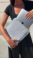 A woman in a black dress holds a light blue and gray chunky knit laptop sleeve containing a silver laptop, set against a light gray outdoor wall.