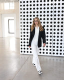 Woman in a black jacket and white pants standing in front of a black and white patterned wall.